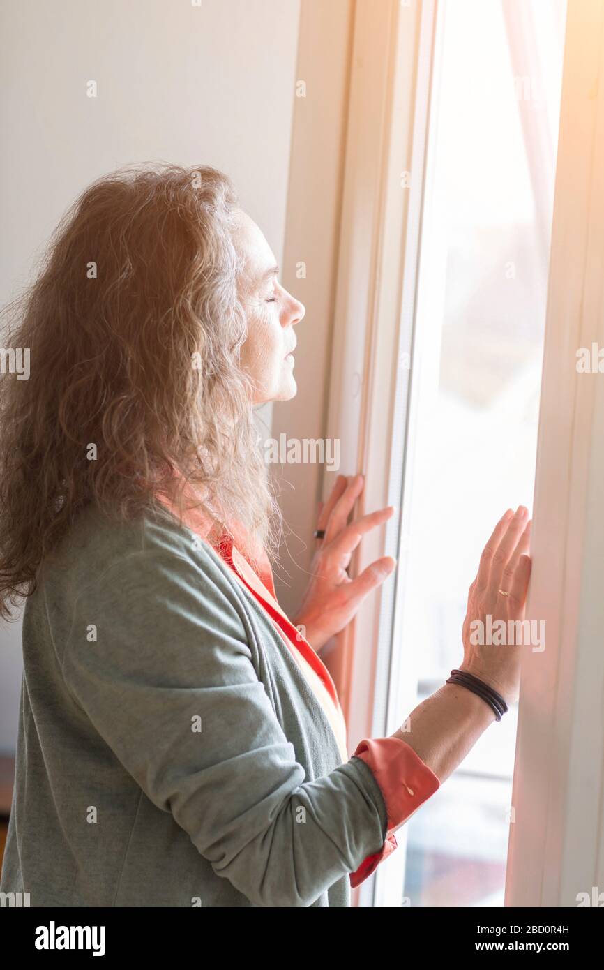 Attractive middle-aged woman enjoying the warmth of the sunlight as she stands in front of a window with closed eyes and a blissful expression Stock Photo