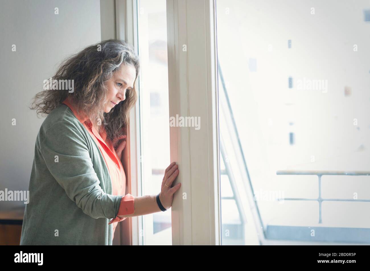 Attractive mature woman with long curly hair standing indoors looking out of a window with a thoughtful expression at something below Stock Photo