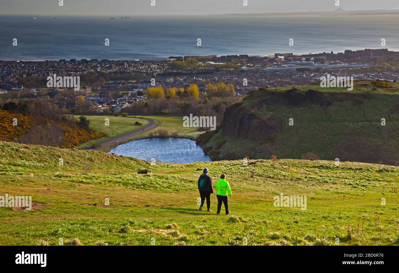 Edinburgh, Scotland, UK. 6th April 2020. As Edinburgh residents prepare to face the third week of the Covid-19 Coronavirus lockdown the morning sun breaks through the previously cloudy sky to give brighter views from Arthur's Seat towards the west over the Salisbury Crags and to the east over Dunsapie Loch towards East  Lothian.Temperature a mild 10 degrees centigrade. Pictured: two female walkers it is assumed taking their permitted daily excercise and enjoying the milder weather on an early morning walk. Credit: Arch White/Alamy Live News. Stock Photo