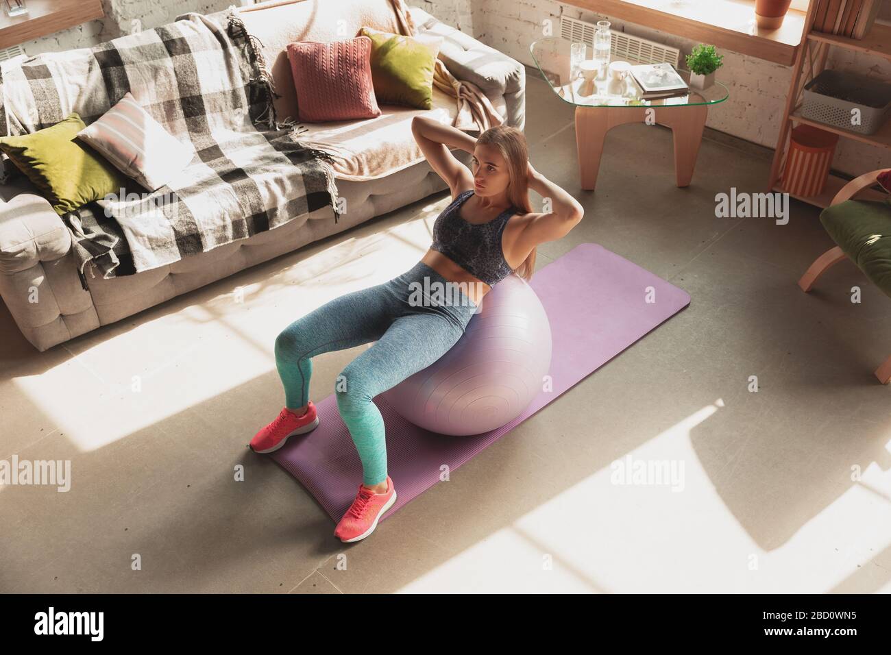 Young woman teaching at home online courses of fitness, aerobic, sporty lifestyle while quarantine. Getting active while isolated, wellness, movement concept. Exercises with fitball for lower body. Stock Photo