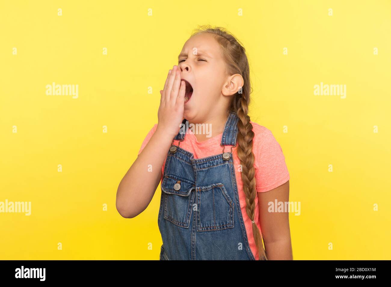Portrait of exhausted child, drowsy little girl with braid in denim overalls yawning, feeling fatigued, sleep problems concept and lack of energy. ind Stock Photo