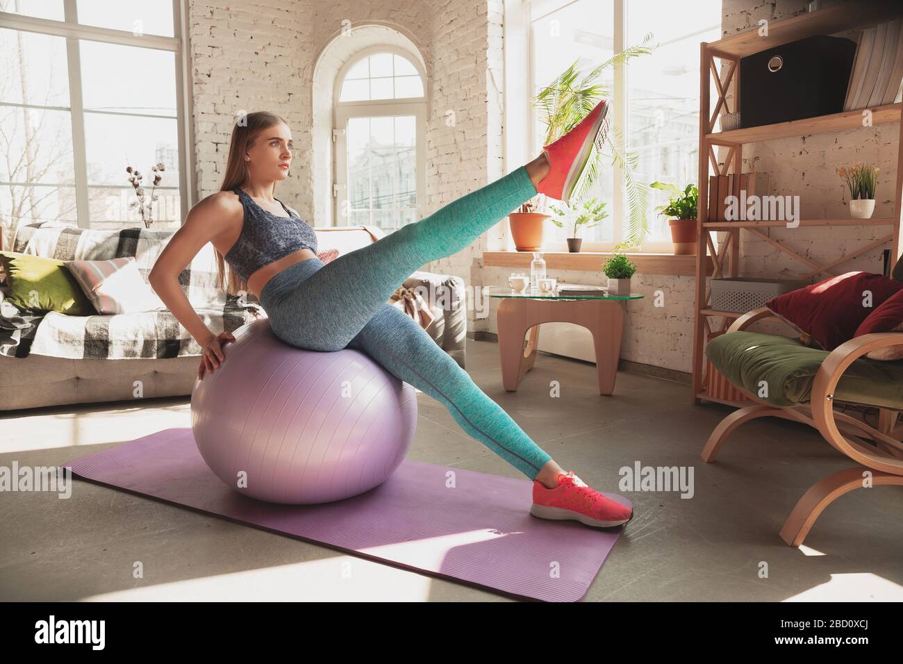 Young woman teaching at home online courses of fitness, aerobic, sporty lifestyle while quarantine. Getting active while isolated, wellness, movement concept. Exercises with fitball for lower body. Stock Photo