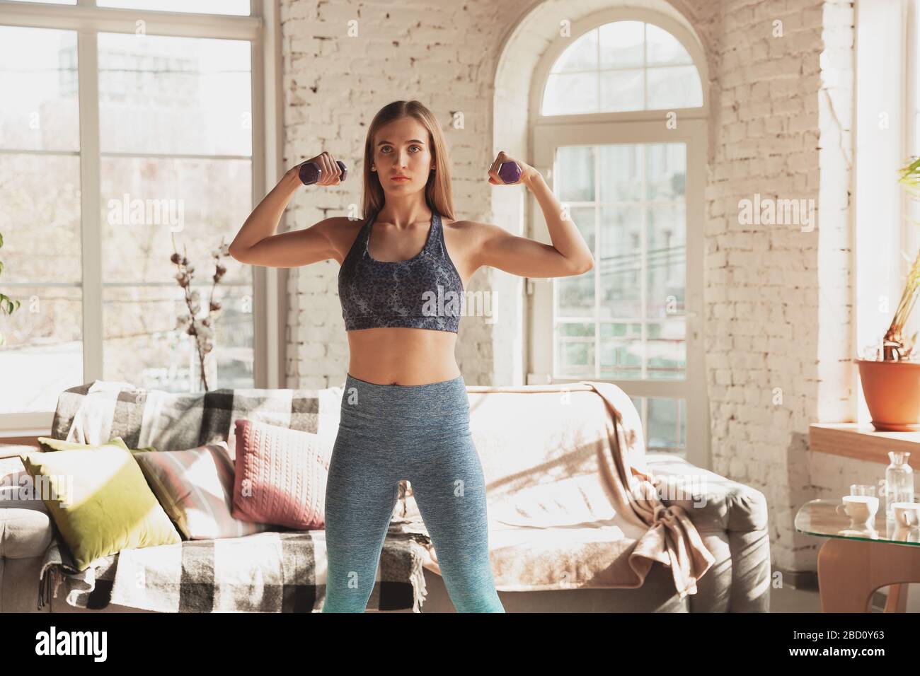 Young woman teaching at home online courses of fitness, aerobic, sporty lifestyle while quarantine. Getting active while isolated, wellness, movement concept. Exercises with weights, balance. Stock Photo