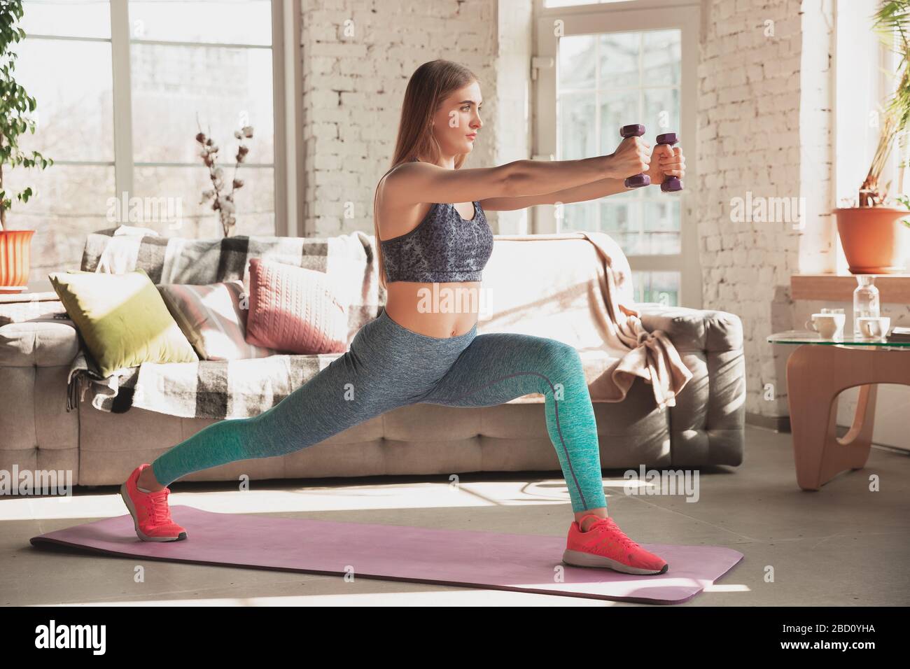 Young woman teaching at home online courses of fitness, aerobic, sporty lifestyle while quarantine. Getting active while isolated, wellness, movement concept. Exercises with weights, balance. Stock Photo