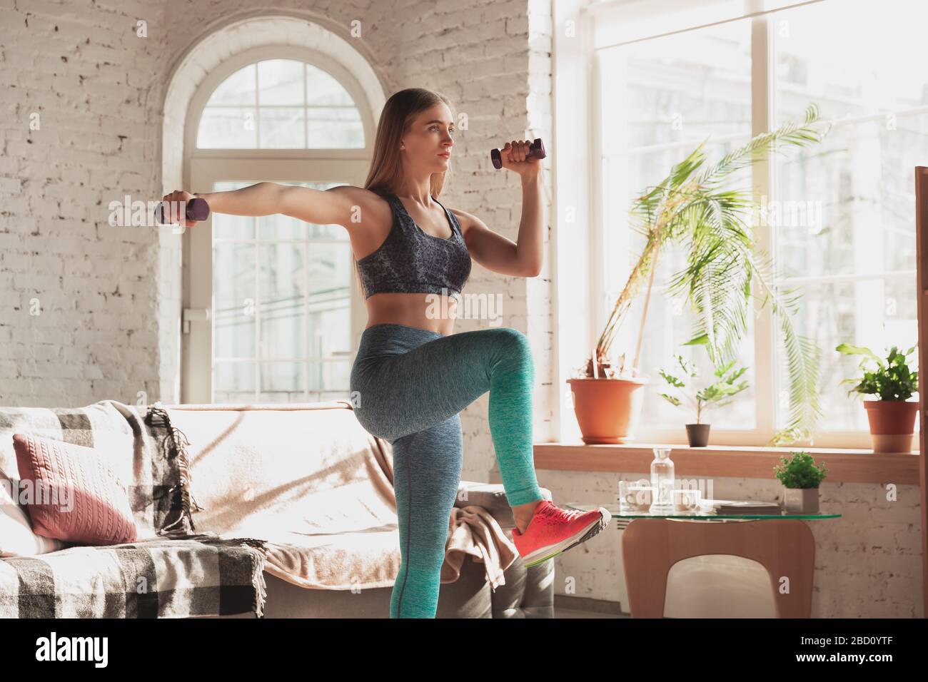 Young woman teaching at home online courses of fitness, aerobic, sporty lifestyle while quarantine. Getting active while isolated, wellness, movement concept. Exercises with weights, balance. Stock Photo