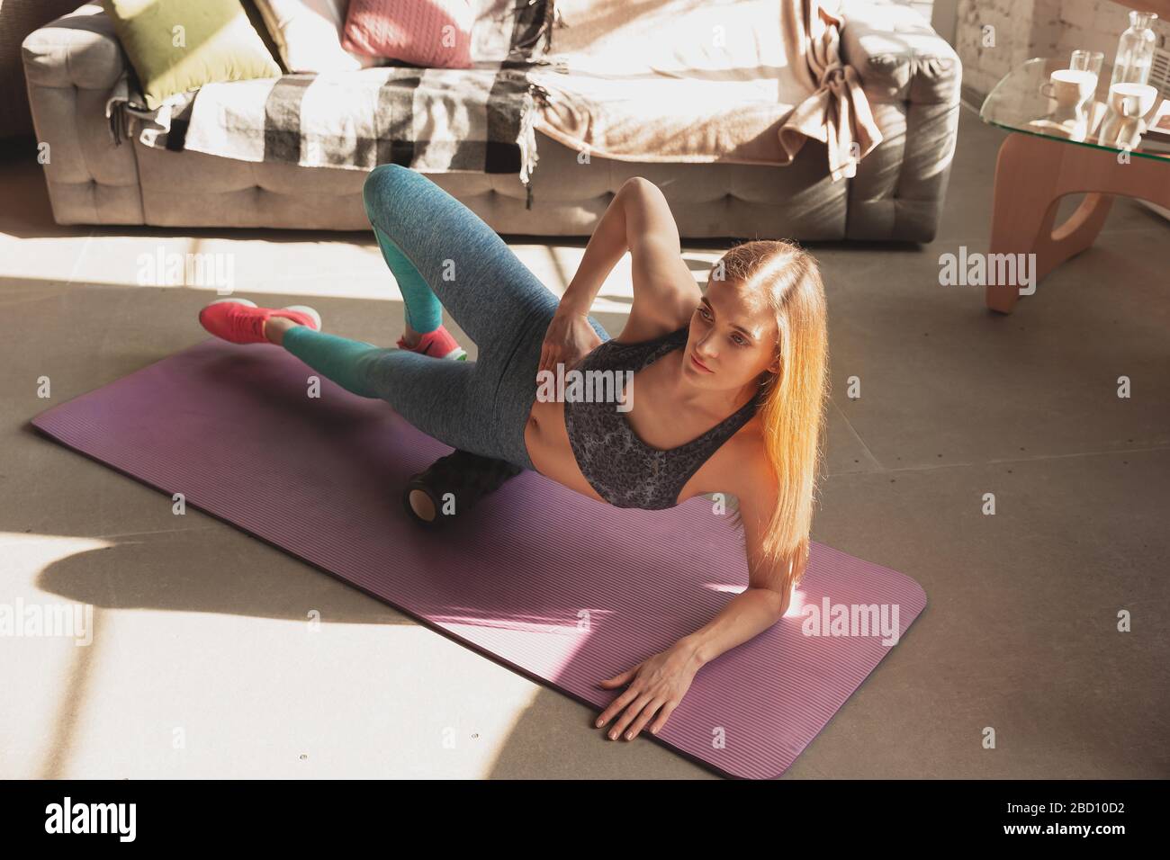Young woman teaching at home online courses of fitness, aerobic, sporty lifestyle while quarantine. Getting active while isolated, wellness, movement concept. Exercises with roller, balance. Stock Photo
