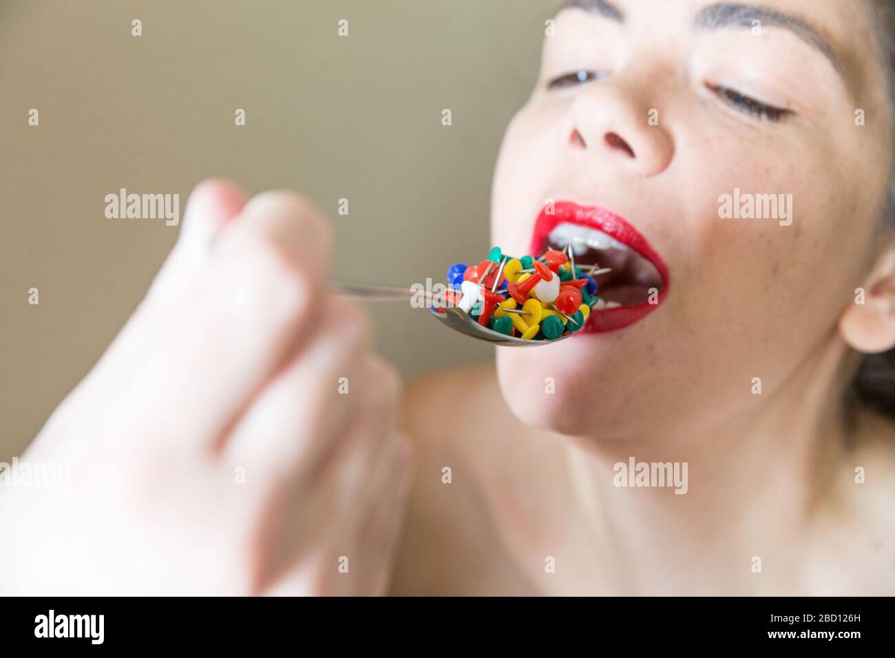 Beautiful girl with red lipstick eating colored sharp drawing pins from a  metallic spoon. Conceptual image Stock Photo - Alamy