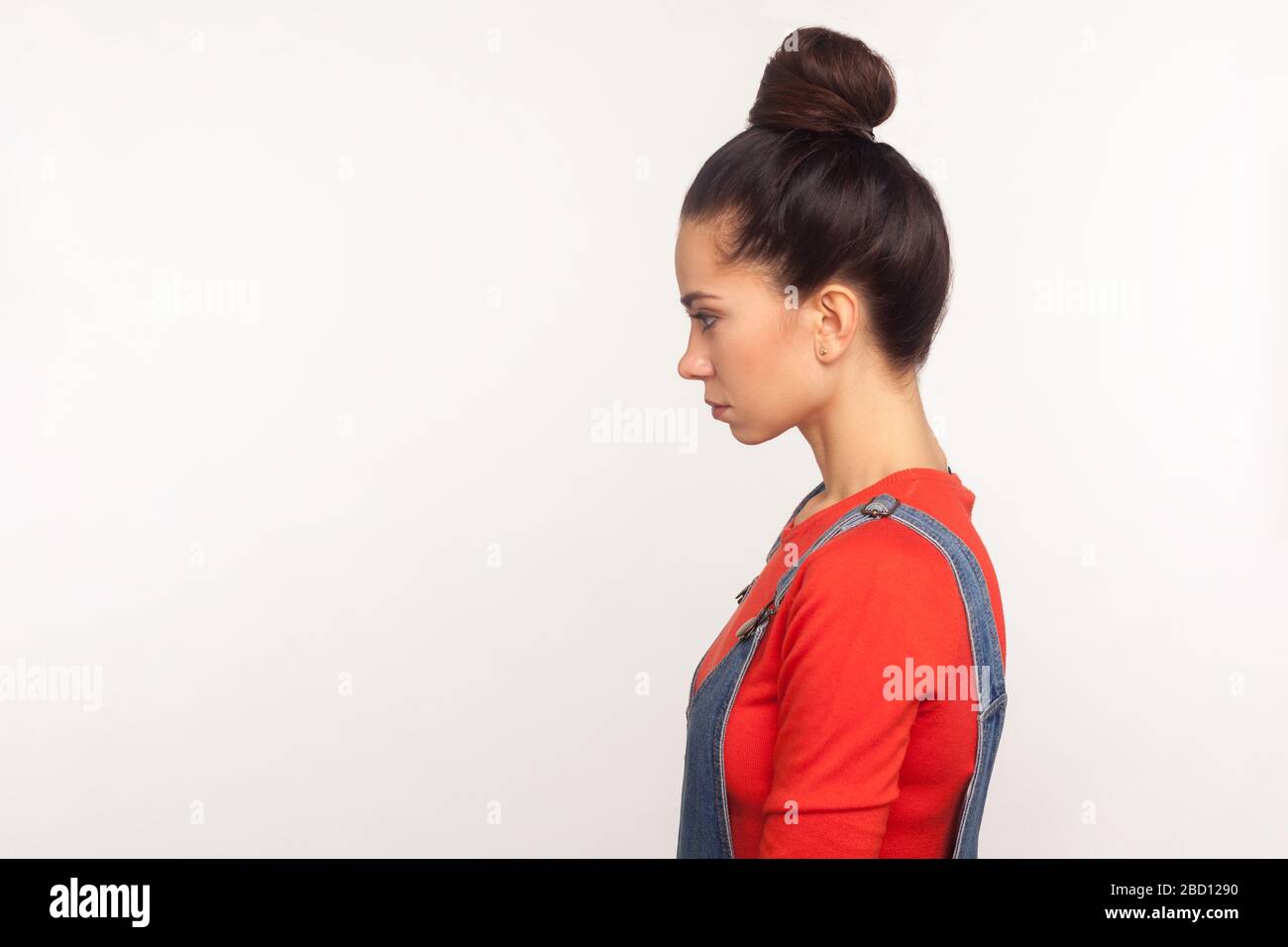 Profile of upset depressed girl with hair bun in denim overalls standing with head down and unhappy look, expressing sadness, worrying about problems. Stock Photo