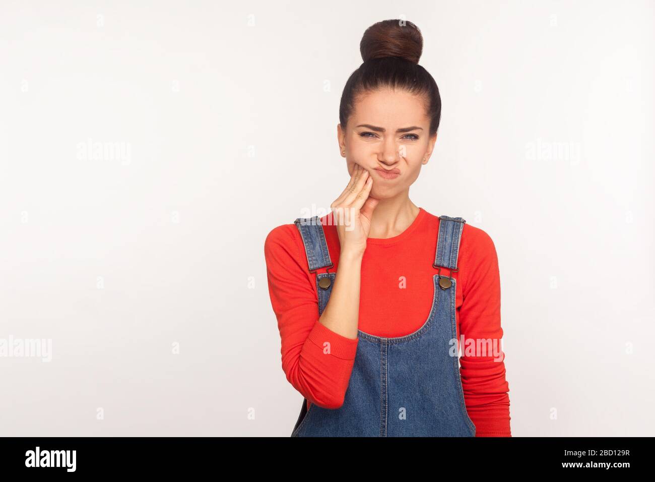 Dental health problems. Portrait of unhealthy girl with hair bun in denim  overalls touching sore cheek, suffering toothache, cavities or gum disease  Stock Photo - Alamy