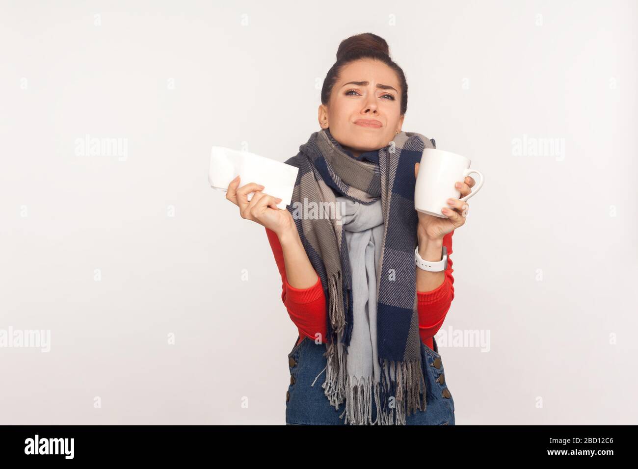 Flu treatment. Depressed unhealthy young woman wrapped in warm scarf holding napkin and tea cup, shivering from cold, suffering fever, high temperatur Stock Photo