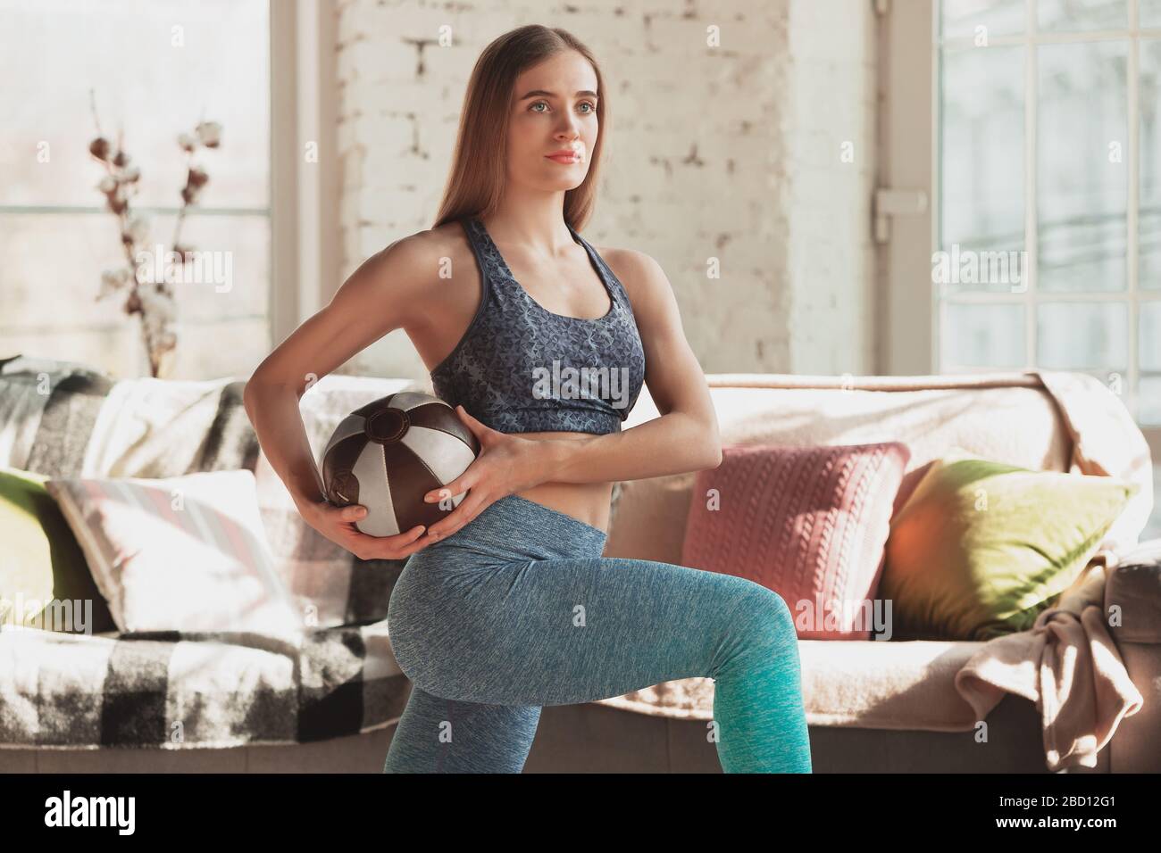Young woman teaching at home online courses of fitness, aerobic, sporty lifestyle while quarantine. Getting active while isolated, wellness, movement concept. Exercises with ball for lower, upper body. Stock Photo