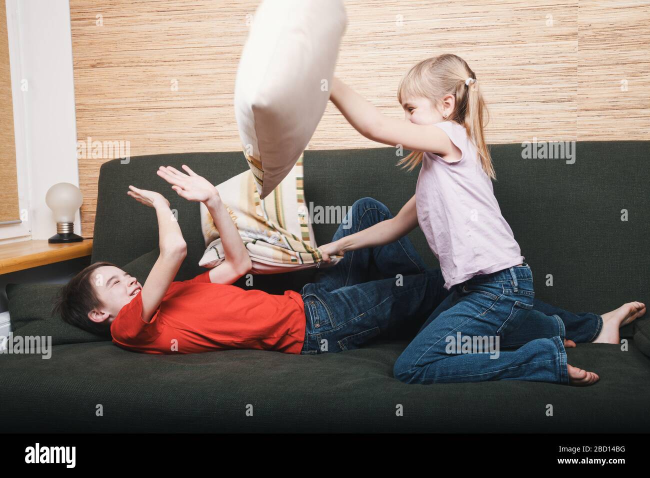 Siblings going mad stuck at home being in self-isolation. Children fighting with pillows on a couch. Quarantine and lockdown protective measures again Stock Photo
