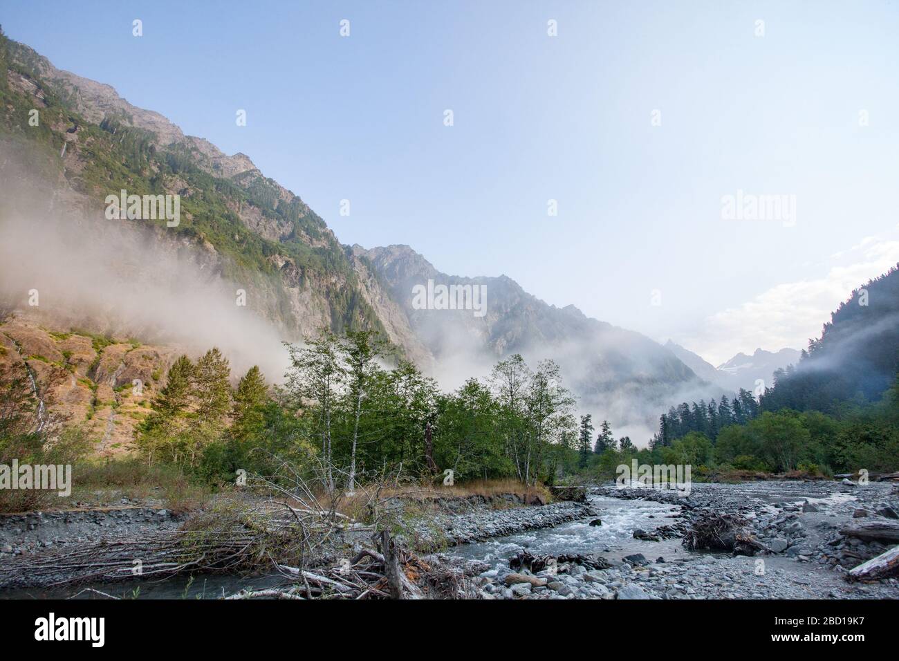 A stream runs through a mountain valley, with low-lying fog and sunlit mountainsides. Stock Photo