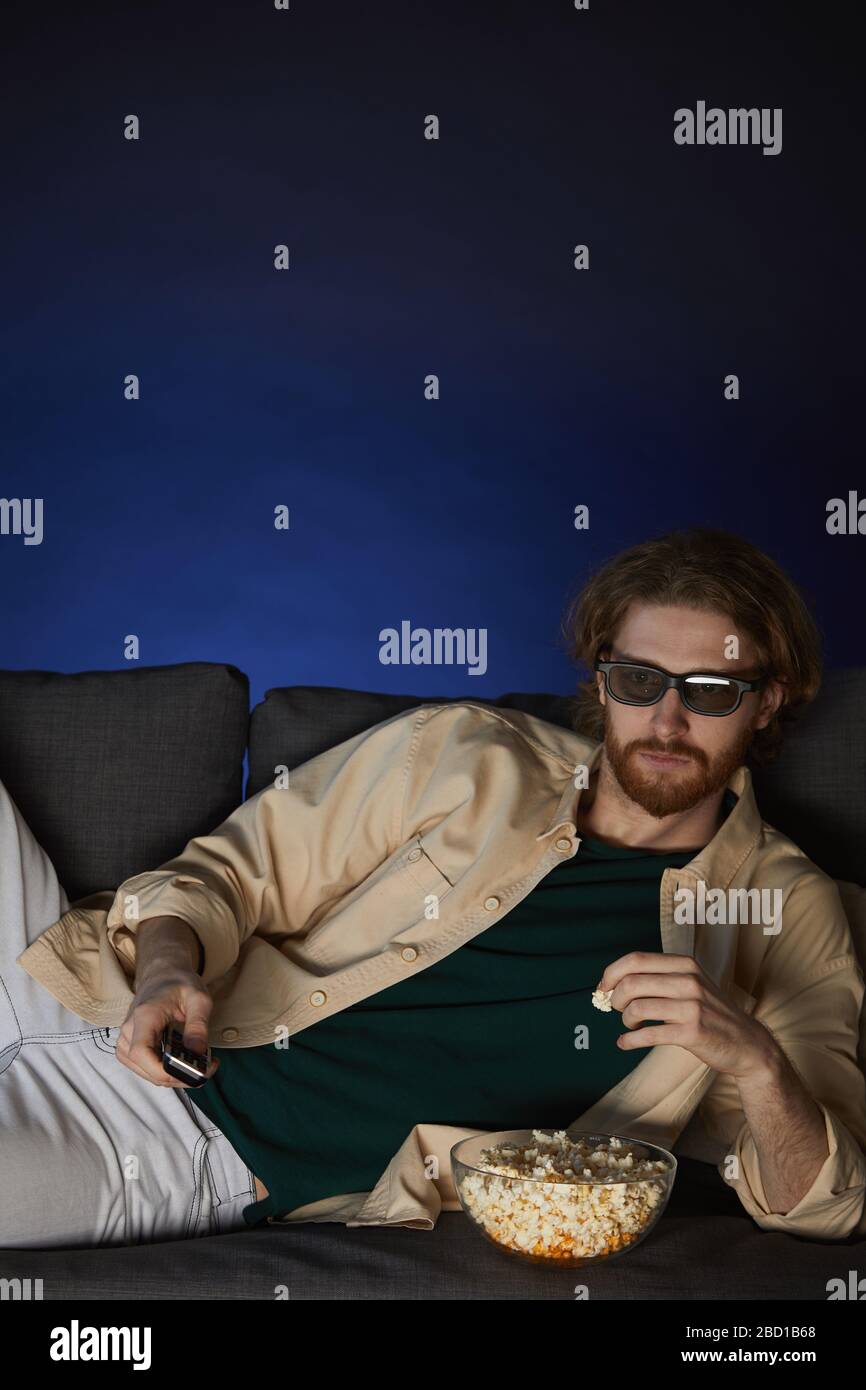 Vertical portrait of modern bearded man watching movie and wearing stereo glasses while lying on couch in dark room, copy space Stock Photo