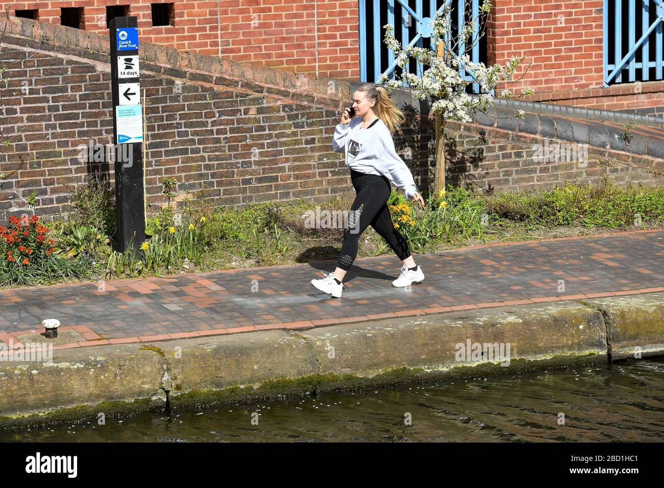 A woman walks by an information sign highlighting preventive measures against Covid19 on a canal in Birmingham city centre, as the Canal and River Trust has started putting up signs warning people to limit use of canal towpaths and have regard for social distancing. Stock Photo