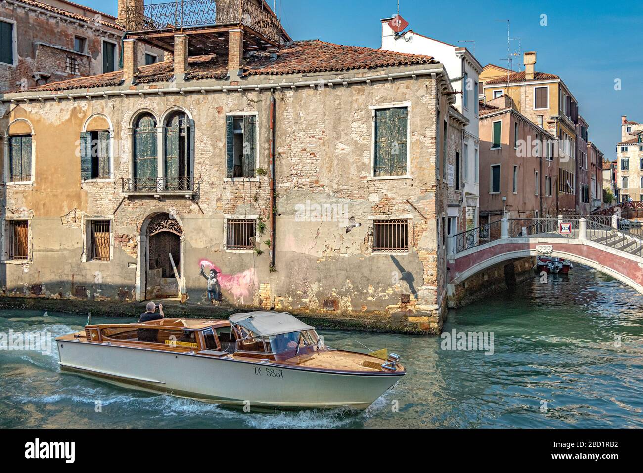 A passenger in a water taxi  takes a picture of the Banksy painting of a child migrant with a pink flare on the wall of the bulding ,Venice ,Italy Stock Photo