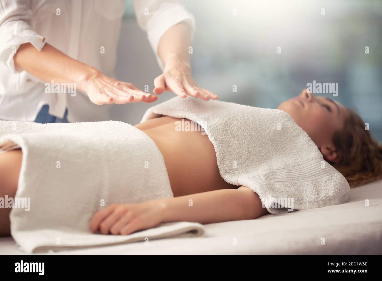 Young girl lying covered with towels over her chest and waist receiving reiki energy through the hands of a therapist. Stock Photo