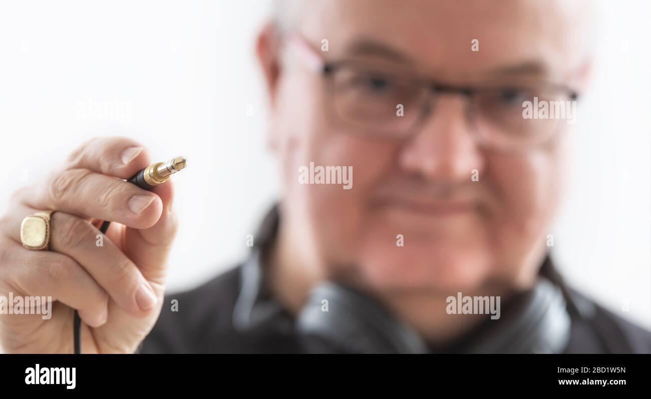 An old man in glasses on white background wearing headphones on his neck with a close-up focus on his hand holding an audio cable jack. Stock Photo