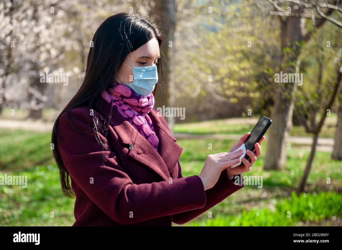 COVID-19. Young Woman in Protective Sterile Medical Face Mask Outdoors Disinfects Phone Antibacterial Napkin. Coronavirus Concept. Stock Photo