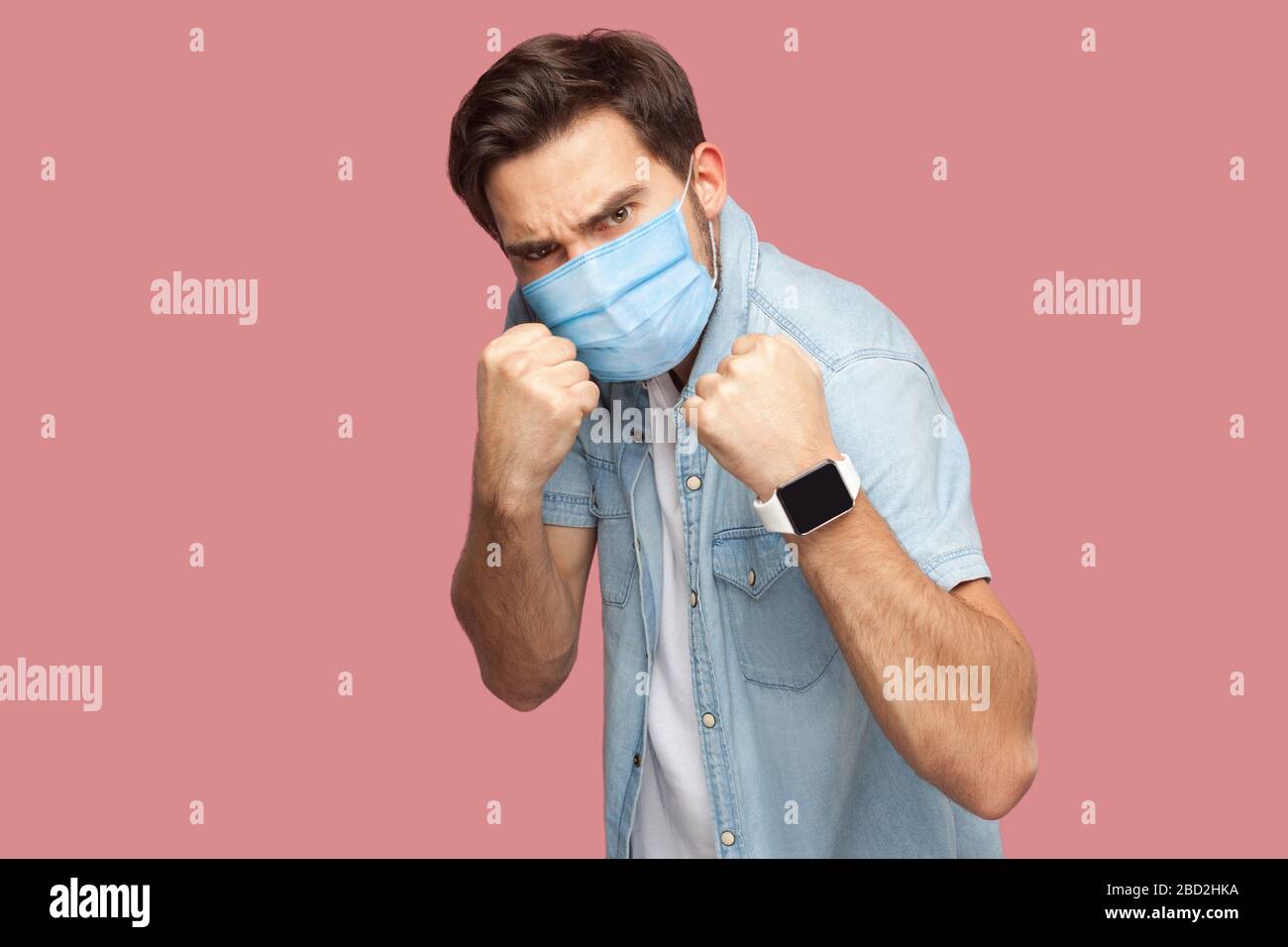 Portrait of angry man with surgical medical mask in blue casual style shirt standing with boxing fists and looking at camera and ready to attack. indo Stock Photo