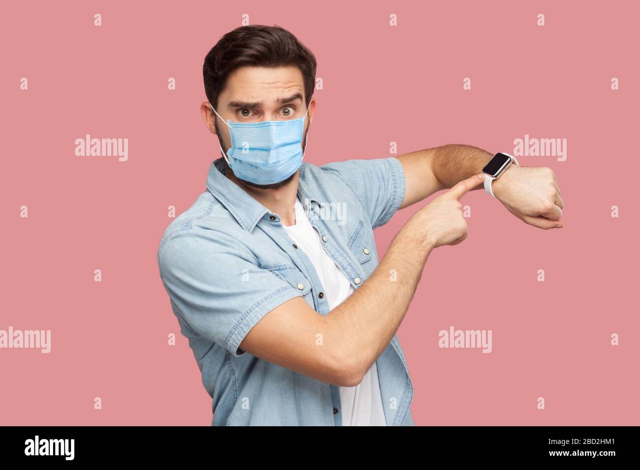 Time is out. Portrait of serious young man with surgical medical mask in blue shirt standing and looking at camera, pointing on his smart watch. indoo Stock Photo