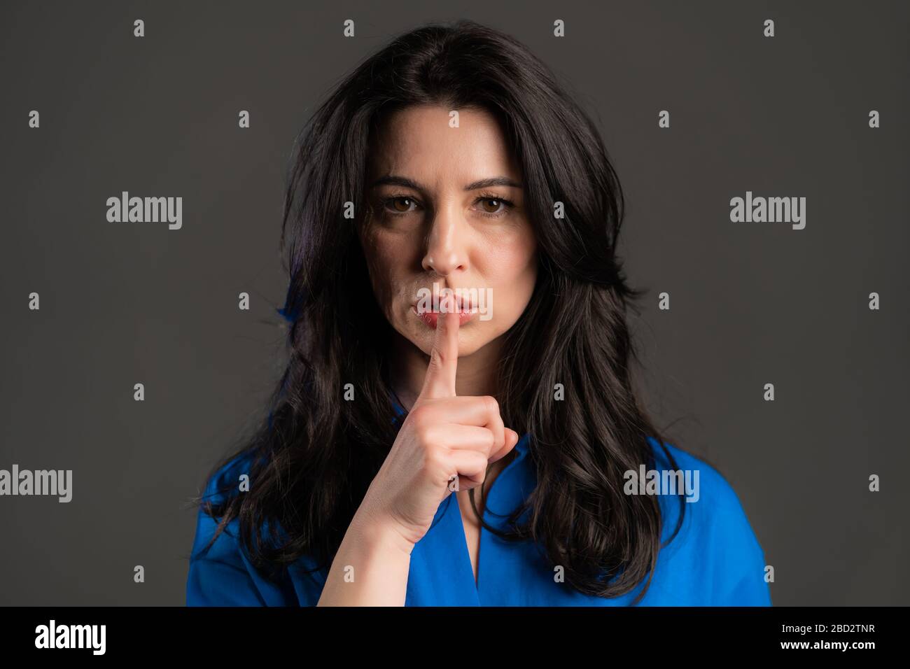 Serious woman with long hair holding finger on her lips over grey background. Gesture of shhh, secret, silence. Close up. Stock Photo