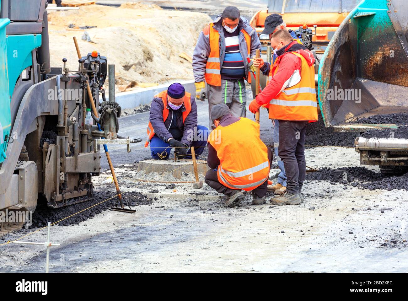 At a new work site, among large pavers, a workers team in protective masks discusses and sets the necessary level for asphalting the road. Stock Photo
