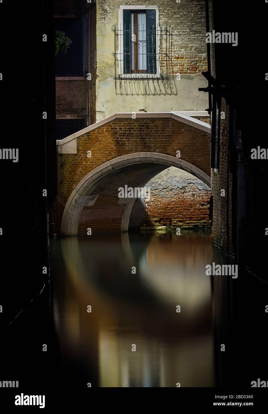 Night shot of a lonely round stone bridge, partially hidden and framed by two dark walls, reflecting on a Venetian canal Stock Photo