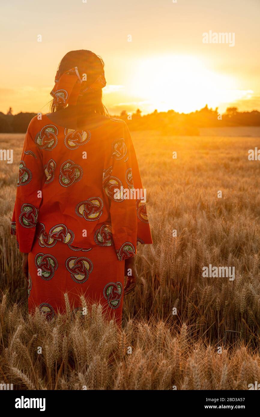African woman in traditional clothes standing in field of barley or wheat crops at sunset or sunrise Stock Photo