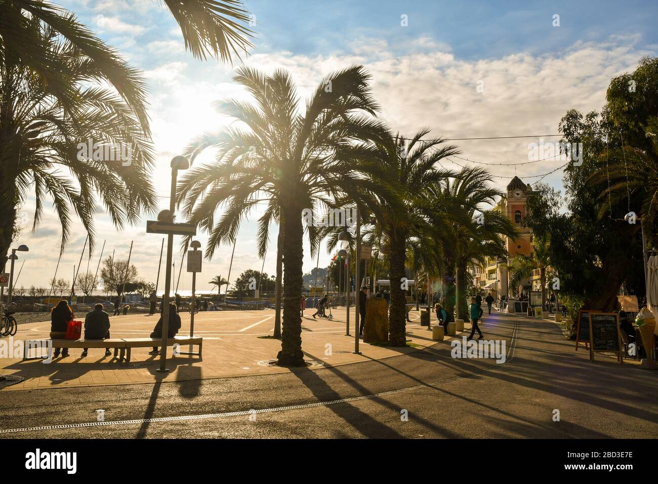 Backlit view of the waterfront of the coastal town of Riviera of Flowers with people walking and sitting on benches, Arma di Taggia, Liguria, Italy Stock Photo