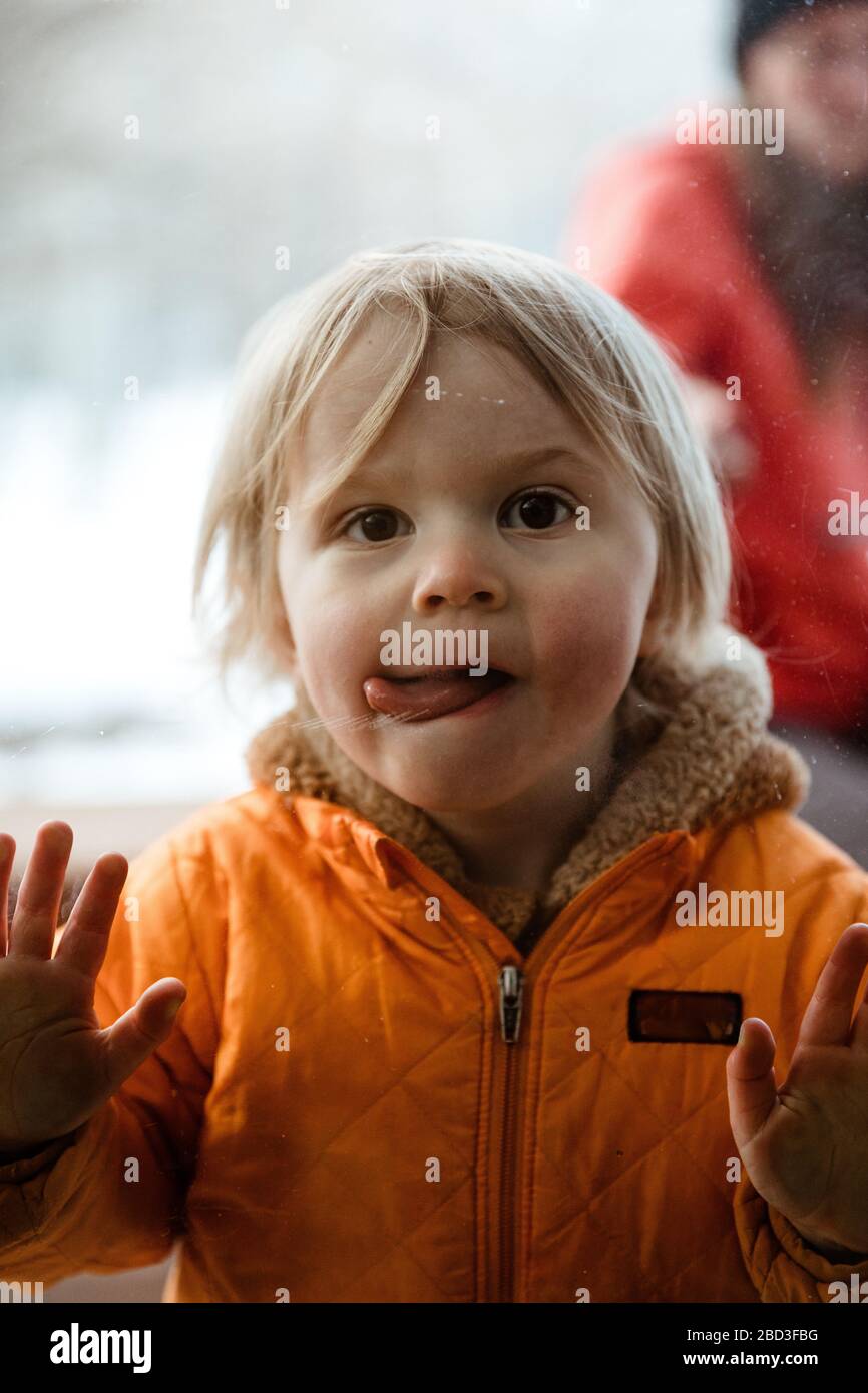 girl toddler pushes on window looks through glass with twisted tongue Stock Photo