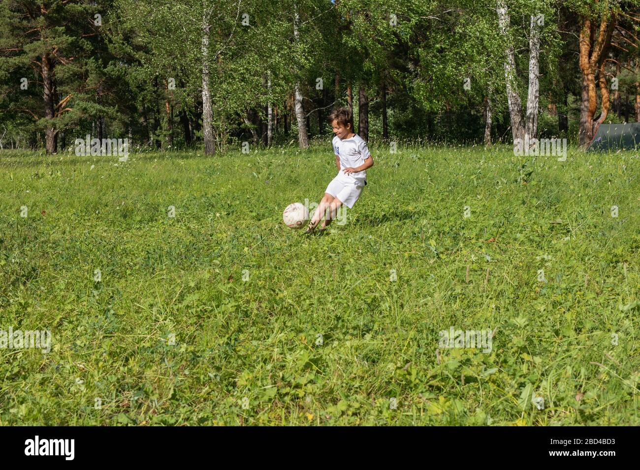 9 years old boy in uniform plays football on green meadow in sunny summer day. child kicks soccer ball, throws it Stock Photo