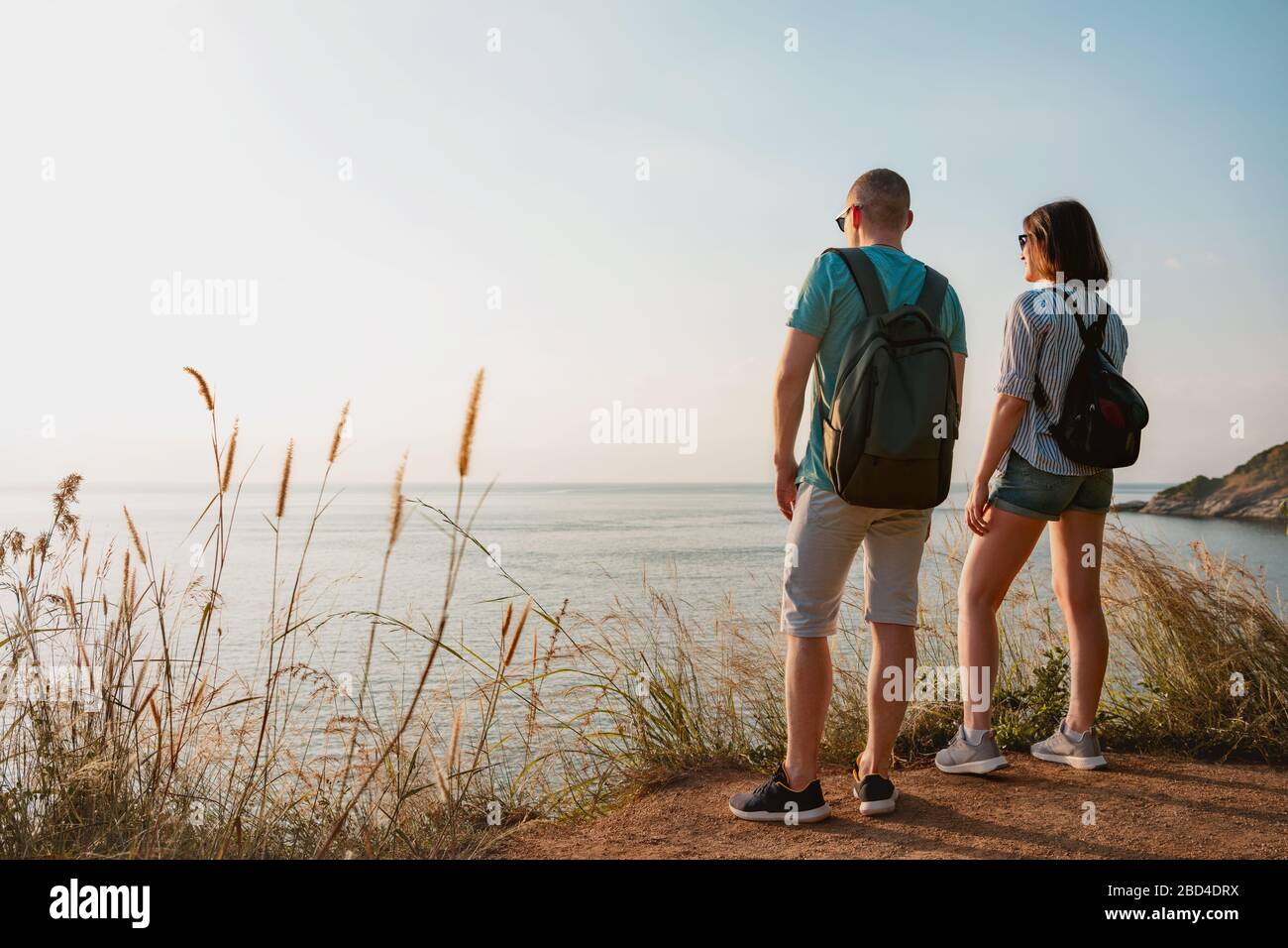 Couple of young hikers with backpacks are standing at sea view point and enjoys sunset Stock Photo
