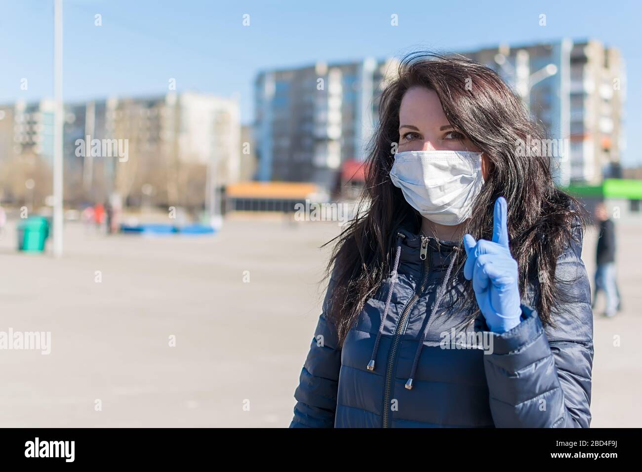 a beautiful girl in a medical anti virus mask warns by pointing her finger up while standing in a city square against the backdrop of shopping centers Stock Photo