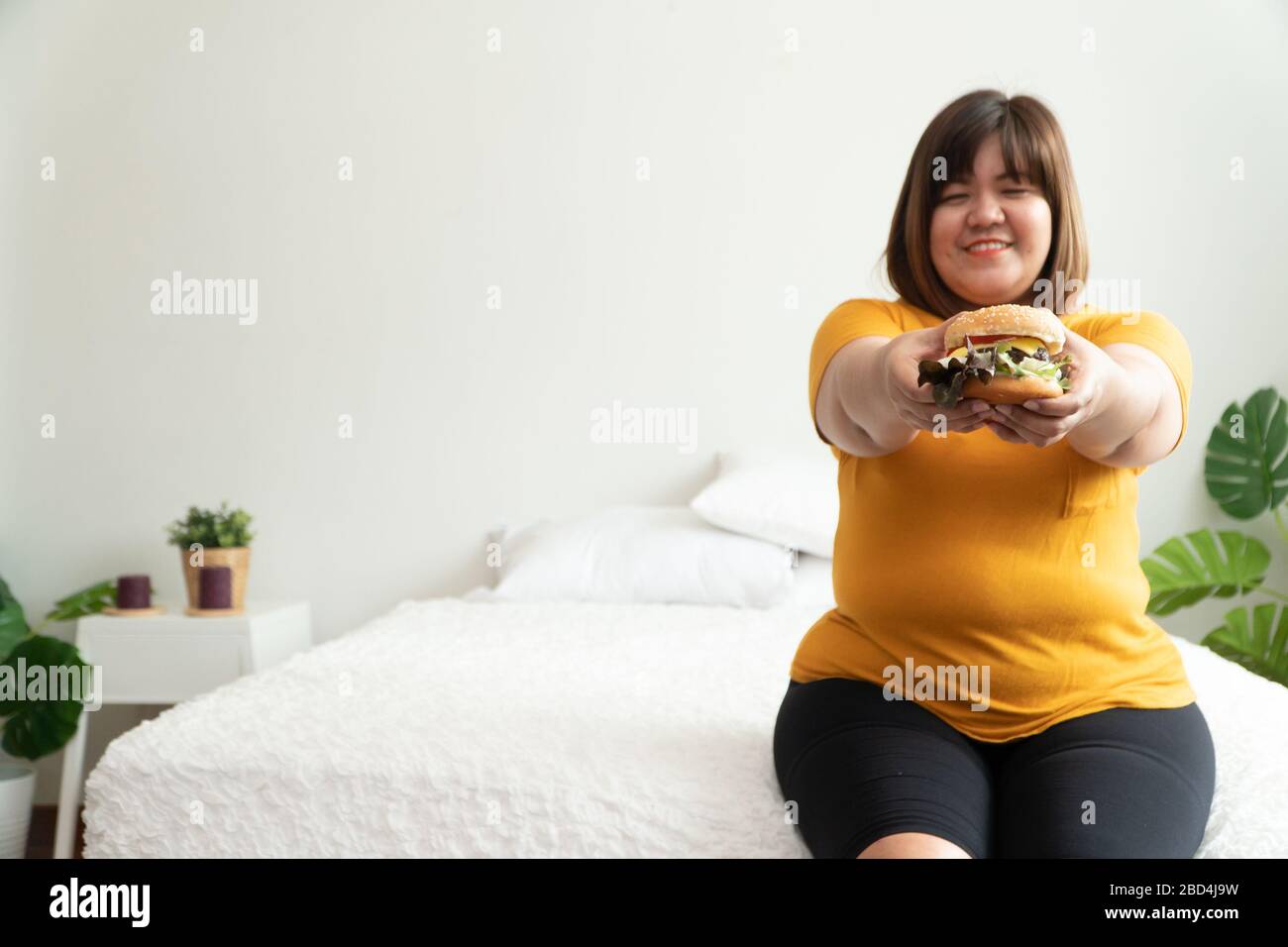 Hungry overweight woman smiling and holding hamburger and sitting in the bedroom, her very happy and enjoy to eat fast food. Concept of binge eating d Stock Photo