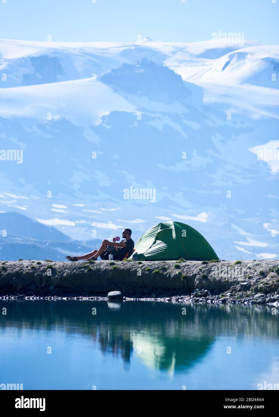 Side view of man enjoying scenery, sitting in chair near tent and drinking hot tea, admiring fantastic view of alpine mountains near lake with fresh clear water. Concept of travelling and camping. Stock Photo