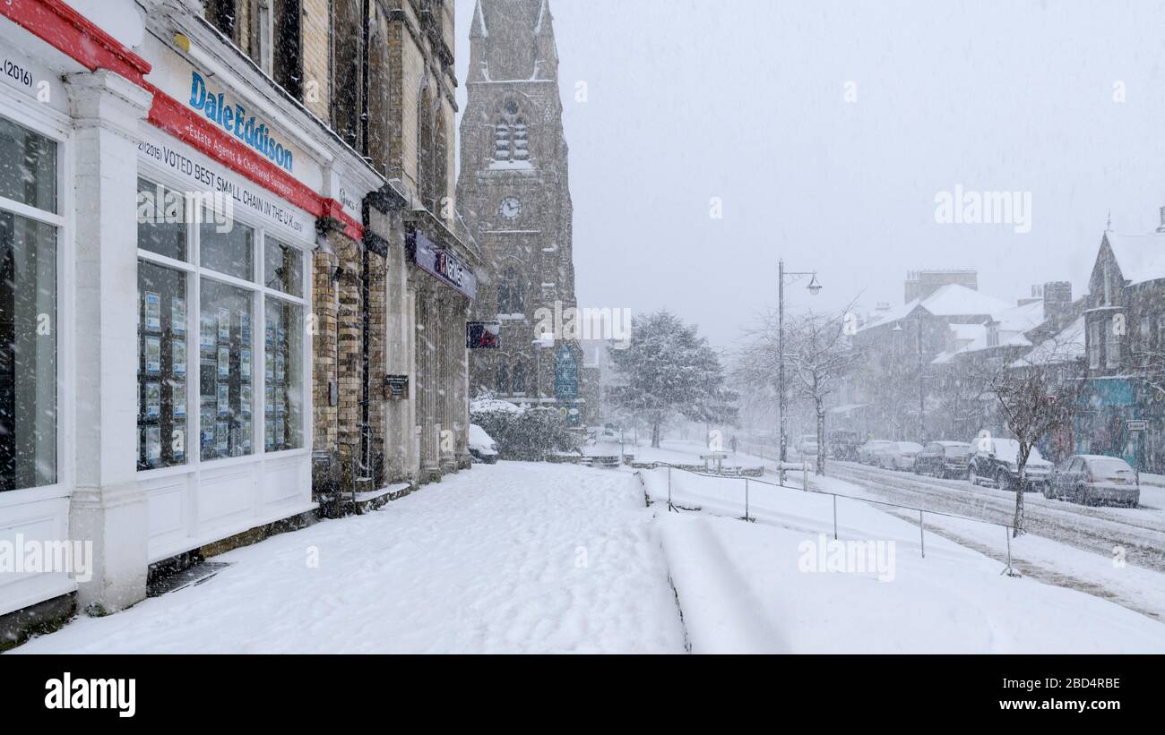 Snowy winter townscape (snowing, snow-covered high street, church, shops & road in scenic town centre) - The Grove, Ilkley, Yorkshire, England, UK. Stock Photo