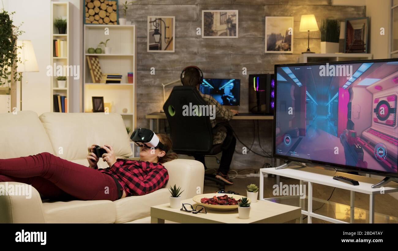 Woman lying on sofa playing video games using vr headset in living room. Boyfriend playing on computer. Stock Photo