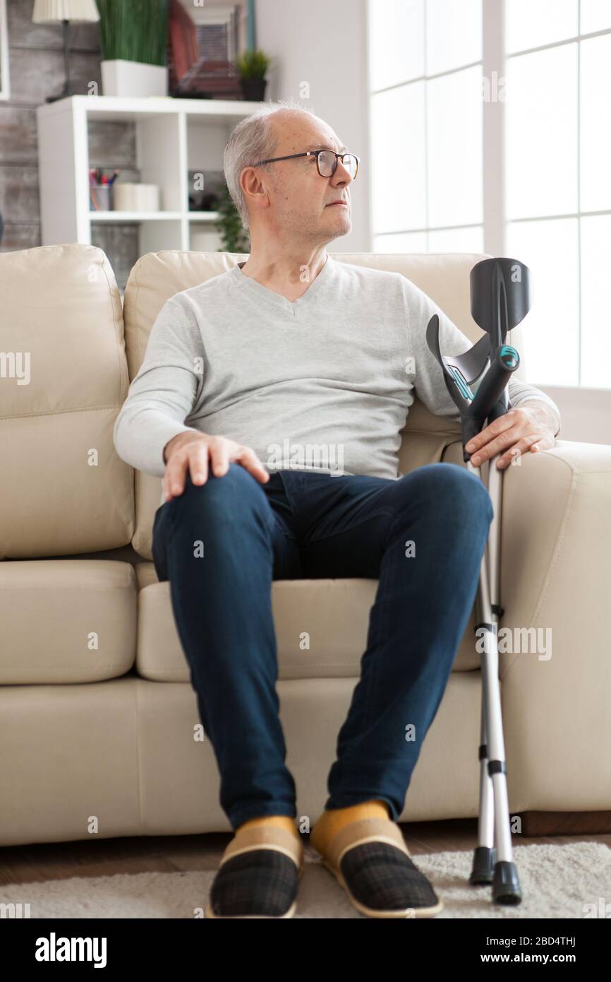 Lonely old man sitting on couch in a nursing home holding crutches and looking away. Stock Photo