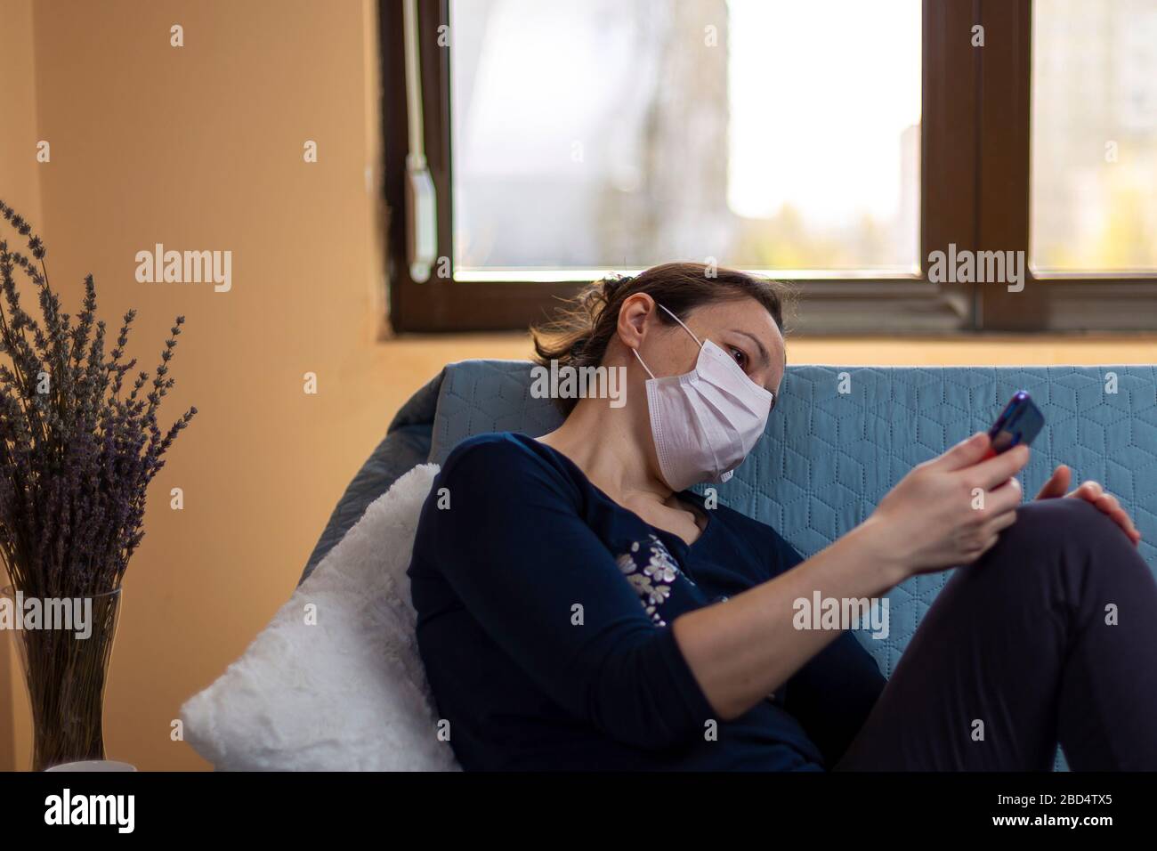 Depressed woman with medical mask sitting on sofa and looking in her smartphone.  Concept of quarantined during the pandemic and feeling the lack of s Stock Photo