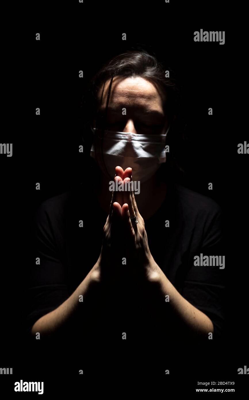 Woman with medical mask standing in prayer in dark background. Concept of infected person standing and praying in a creative light Stock Photo