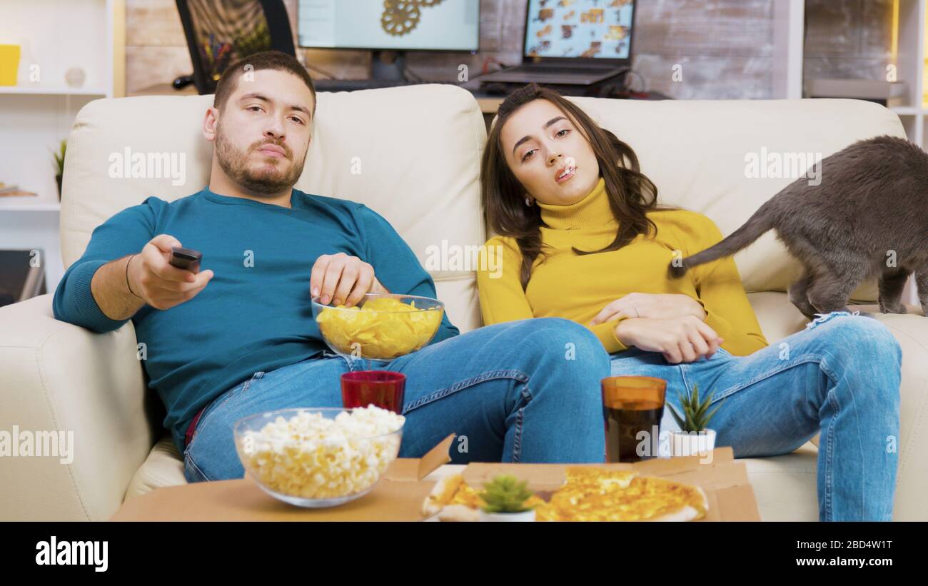 Young couple relaxing on the couch with their cat watching tv. Man eating chips. Stock Photo