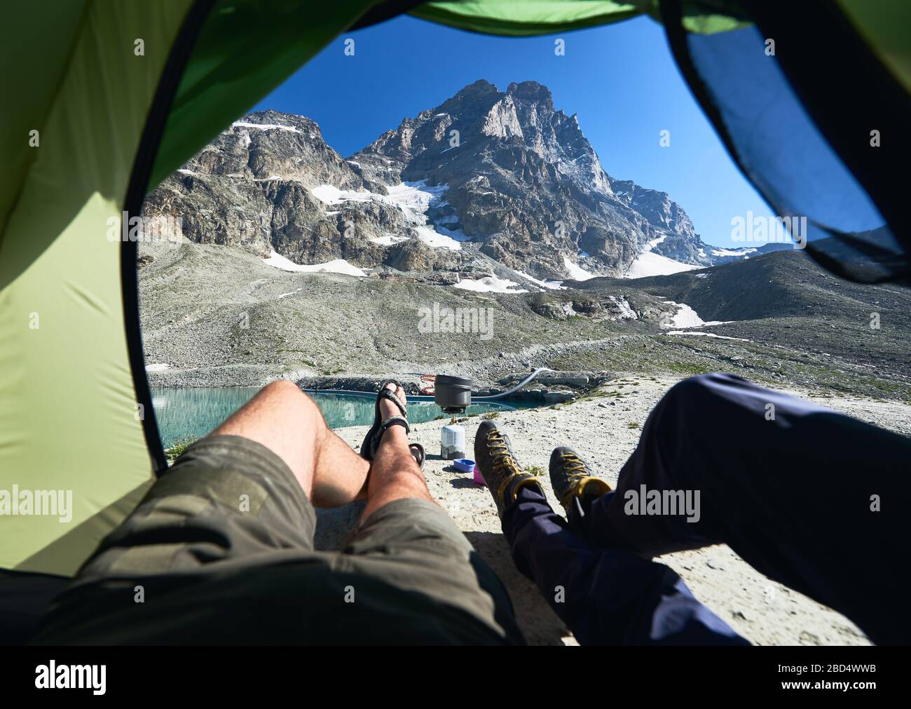 Male legs inside camp tent with snowy mountain on background. Two travelers lying inside tourist tent and enjoying the view of beautiful rocky Alps hills. Concept of travelling, hiking and camping. Stock Photo