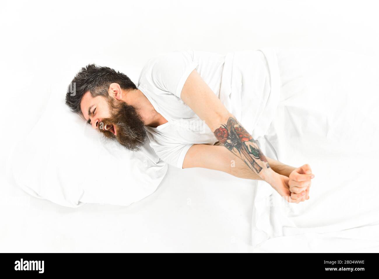 Man with sleepy yawning face stretching while lay in bed, wake up. Man slept well, white background. Good morning concept. Hipster with beard stretching arms, drowsy, sleepy macho, top view. Stock Photo
