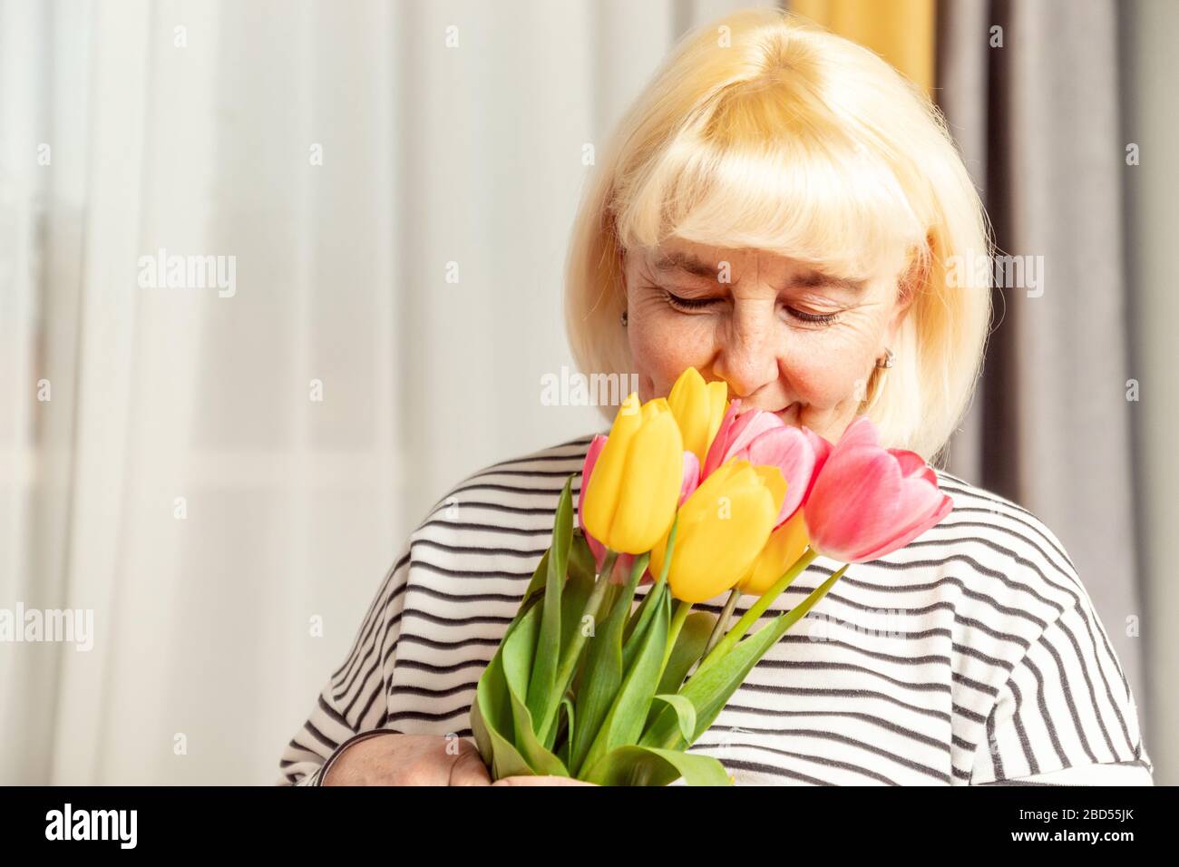 Adult cute blonde woman sniffing a bouquet of tulips flowers on a gray background. Happy mother's day. Stock Photo