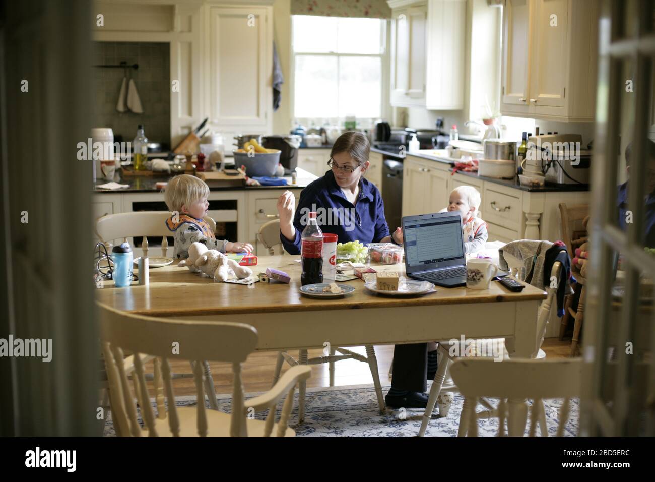 Mother feeding her children dinner at kitchen table whilst working from home during period of self-isolation - 2020 COVID-19 coronavirus pandemic Stock Photo