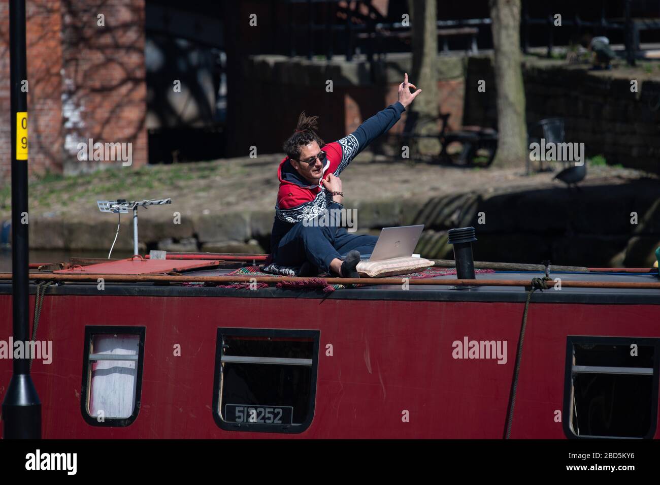 A man uses video conferencing software like Zoom, Hangouts, Skype or Houseparty in the sunshine on top of his canal boat in Castlefield Basin, Manchester city centre, on Tuesday 7th April 2020. Stock Photo