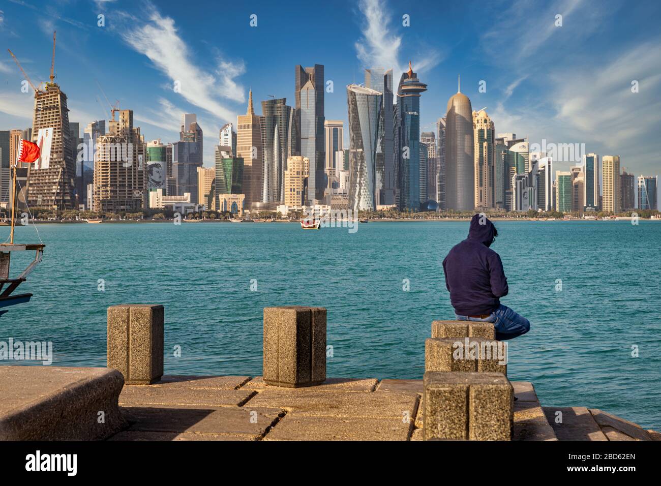 Doha Qatar skyline daylight view with clouds in the sky showing skyscrapers in background and a man sitting on Doha Cor niche in foreground with Qatar Stock Photo