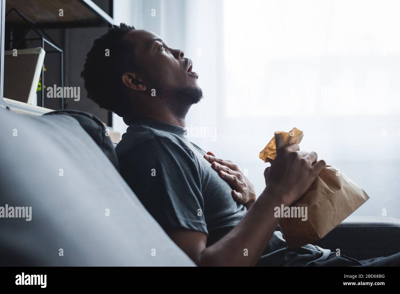 shocked african american man holding paper bag while having panic attack at home Stock Photo