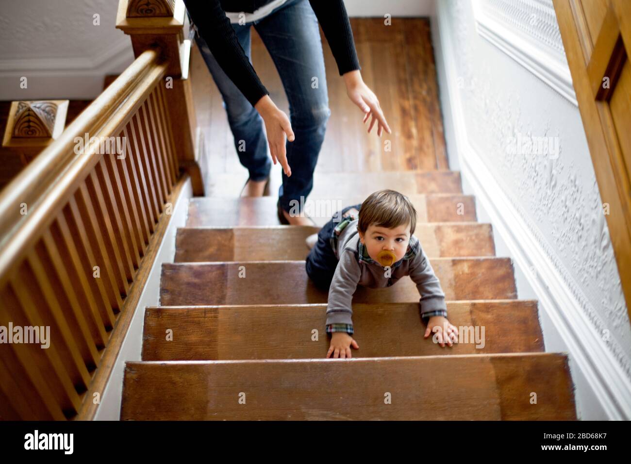 Infant child crawls up a flight of stairs followed closely by his mother. Stock Photo